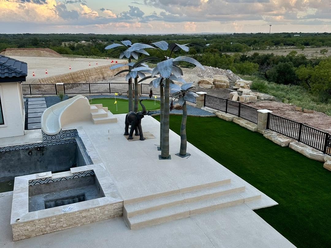 Aerial view of a hilltop backyard with a pool, water slide, putting green, turf, and metal palm trees