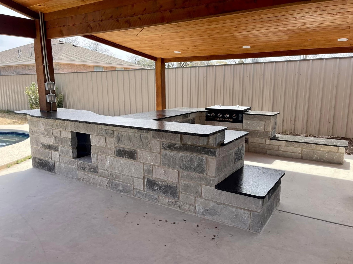 Poolside outdoor kitchen with gray stone base, black granite counters, and a griddle under a wood-ceiling pavilion