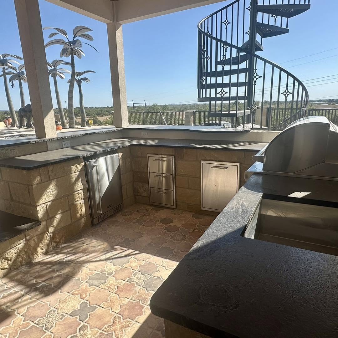 Outdoor kitchen with limestone cabinetry, stainless appliances, and a patterned tile floor beside a spiral staircase