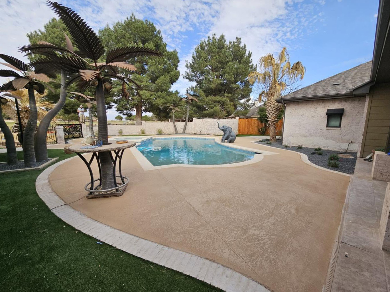 Renovated pool deck in a warm tan finish with sculpted metal palm trees and a turf border