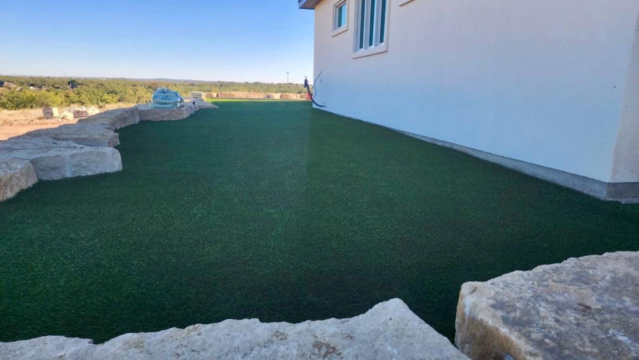 Synthetic turf lawn edged with limestone boulders and a gravel border beside a stone house